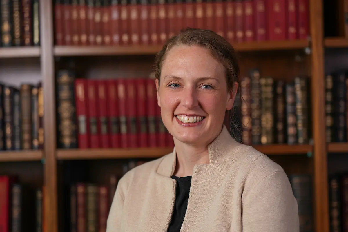 Head and shoulders photo of Ann Jones, smiling and wearing a beige blazer, standing in front of a bookcase.