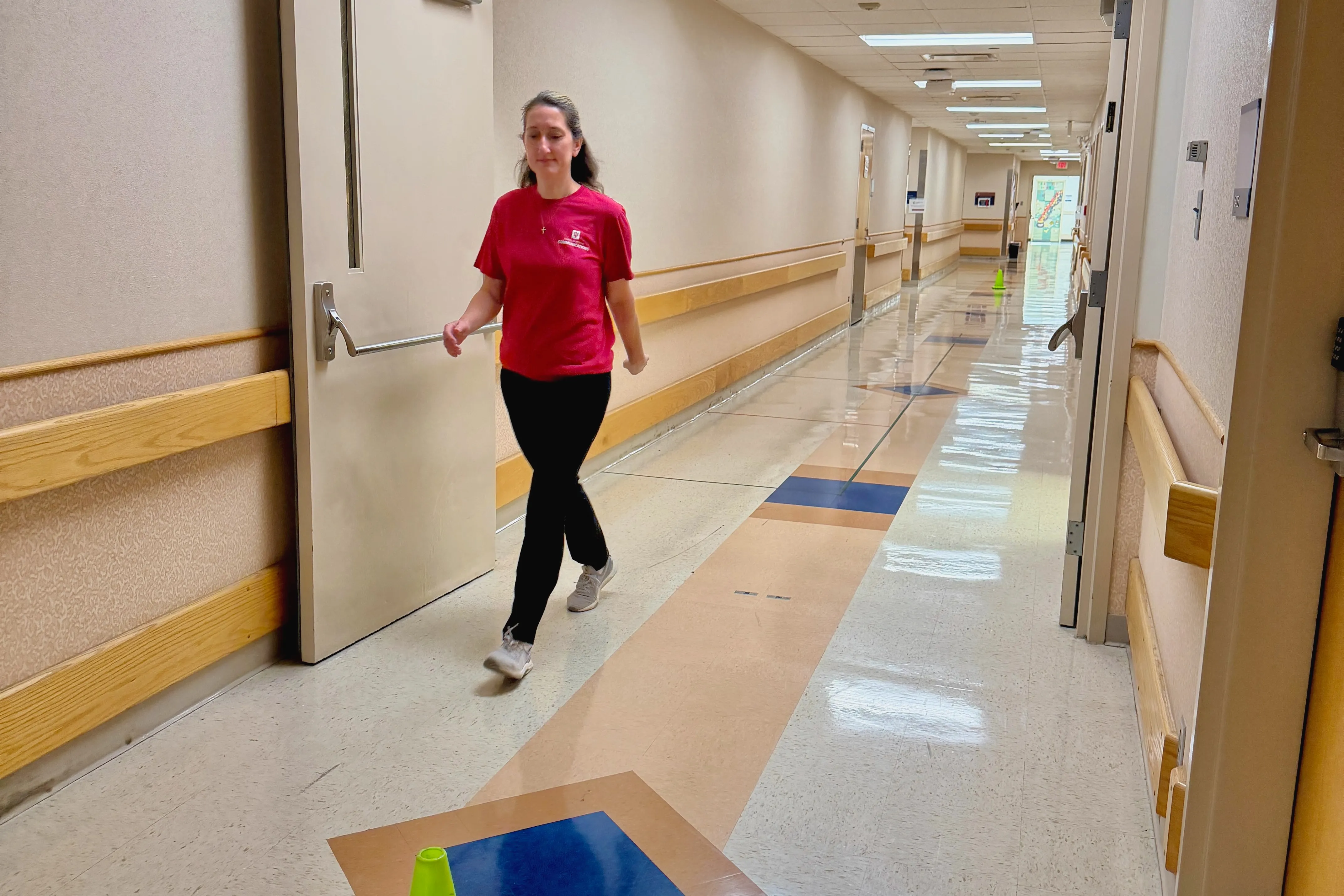A woman in red shirt and black pants walks down a hospital hallway.