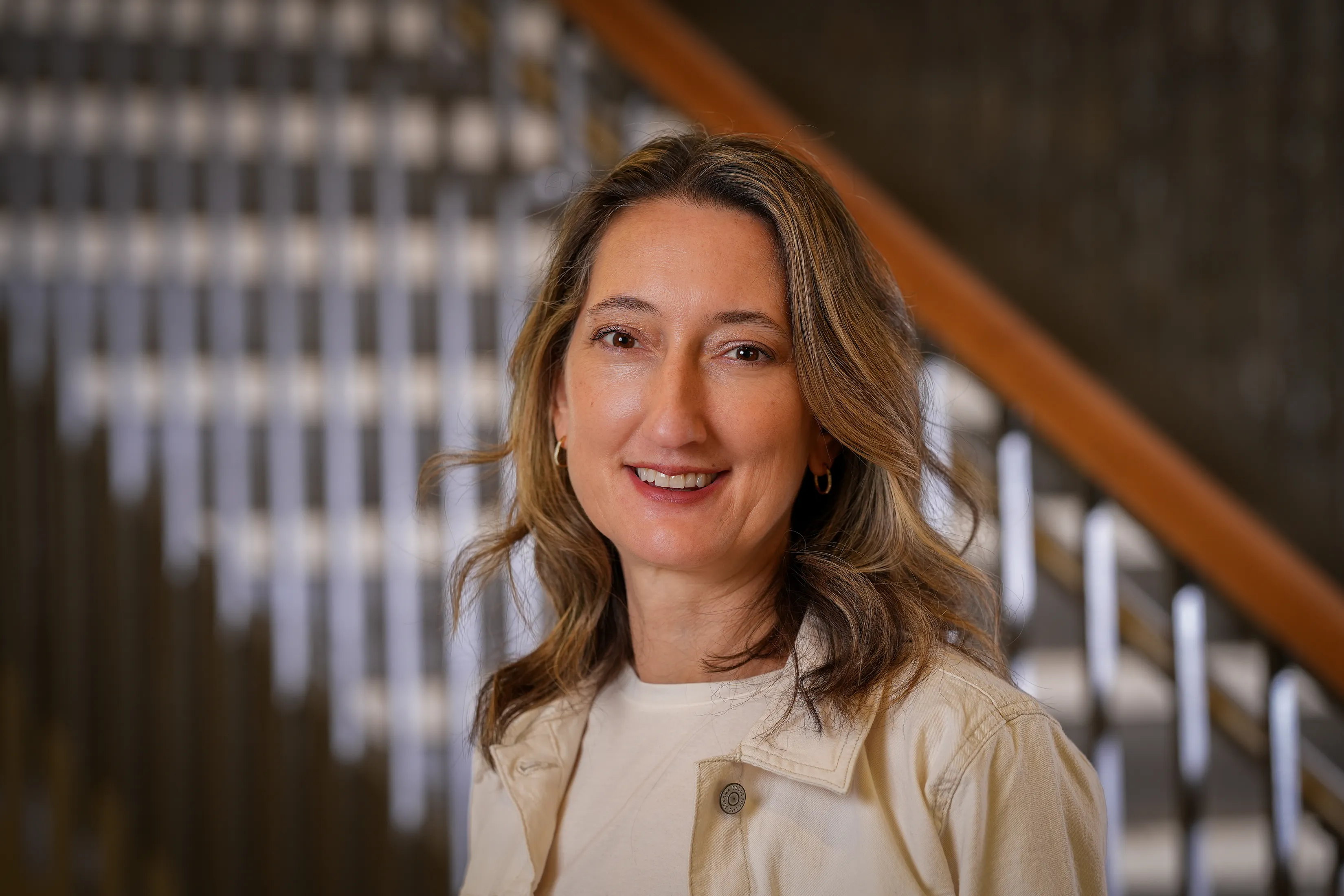 Laura Gates headshot; woman with brown hair wearing a light colored jacket with staircase in the background