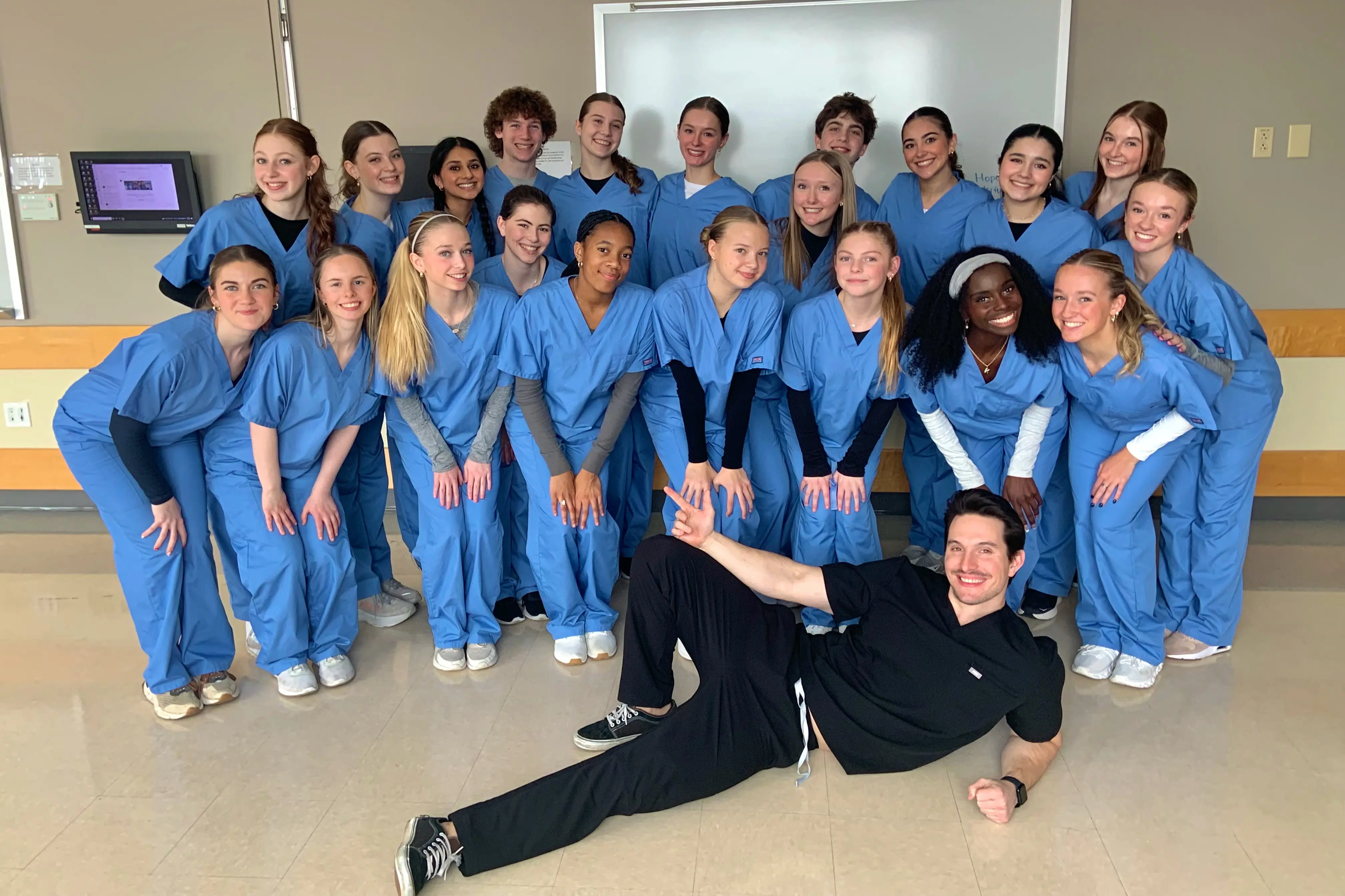 A smiling Male Nurse in black scrubs lies on the floor in front of a group of female nurses in blue scrubs in a hospital setting. 