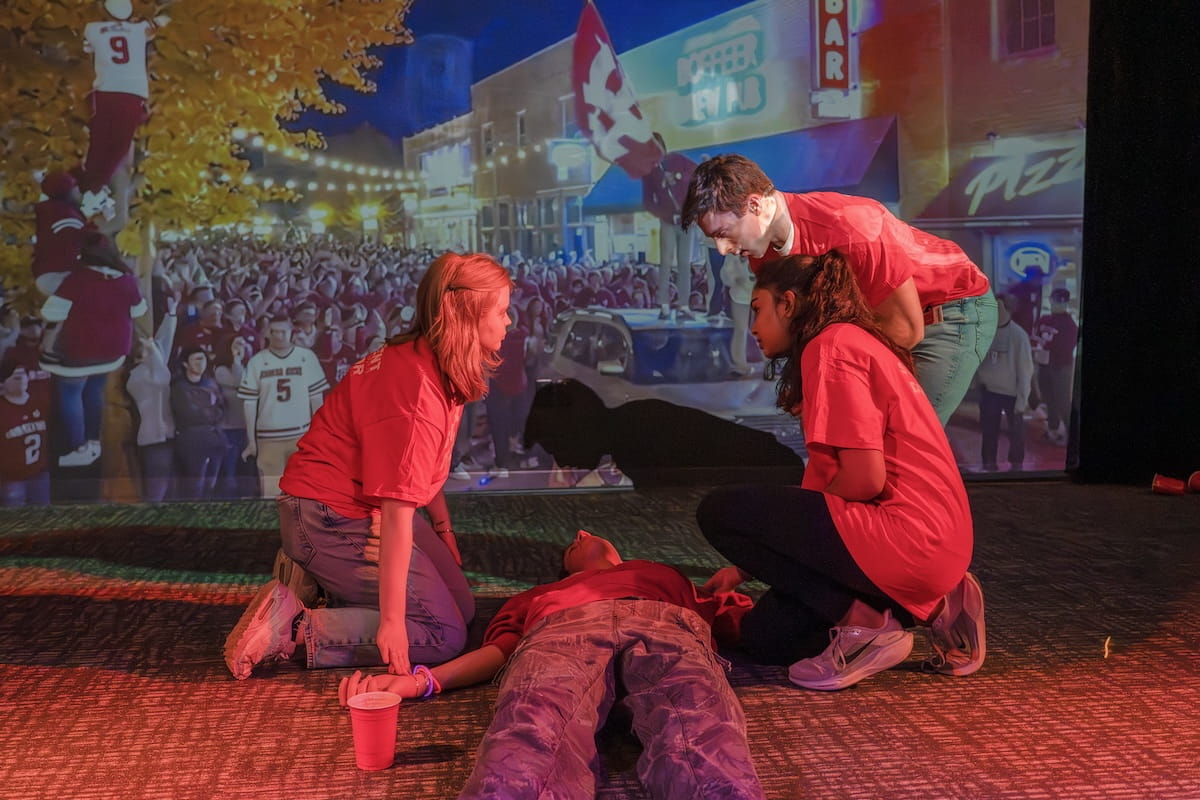 Three medical students in a dimly lit room assess the condition of an actor portraying an overdose during a simulation exercise with video screens behind them