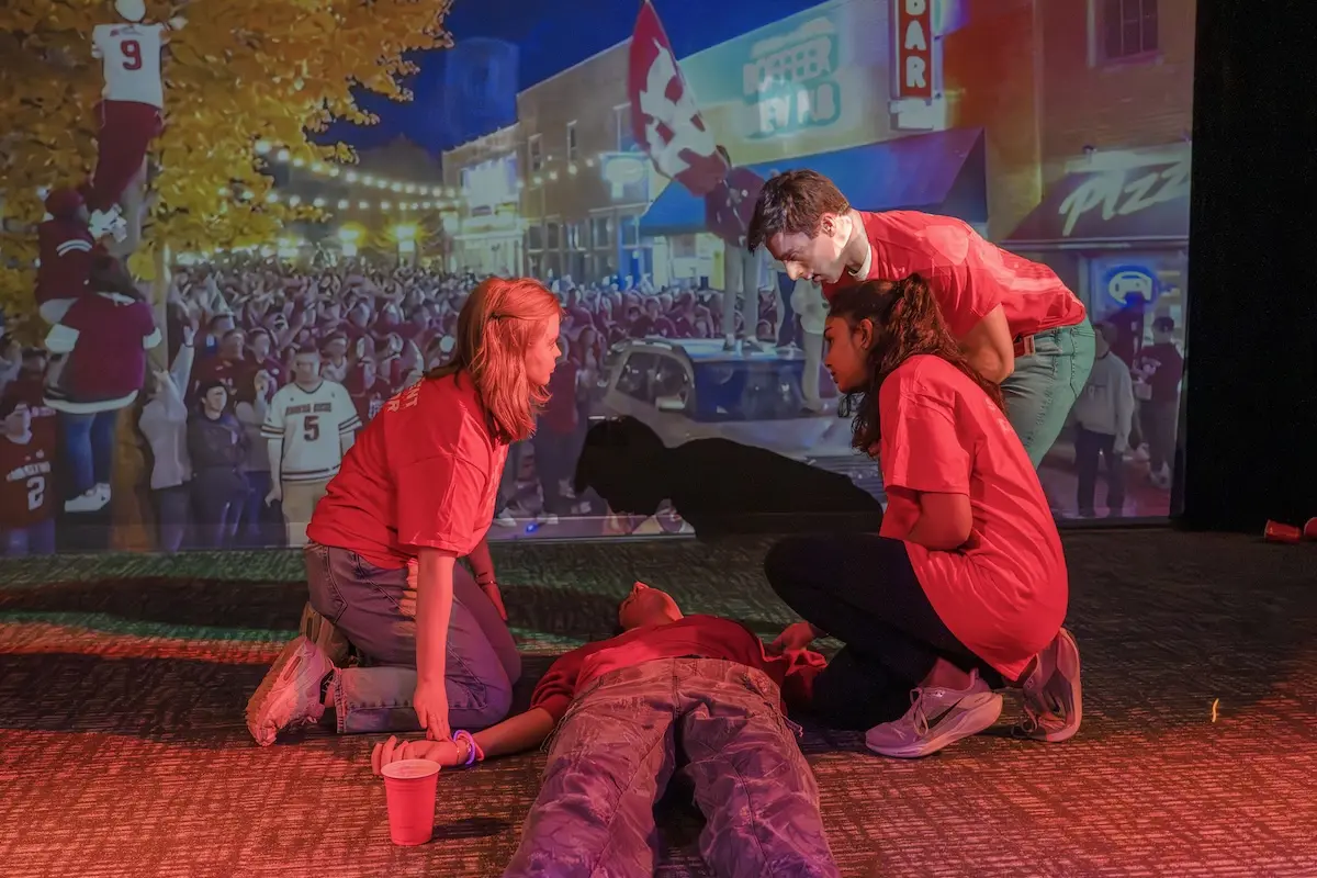 Three medical students in a dimly lit room assess the condition of an actor portraying an overdose during a simulation exercise with video screens behind them