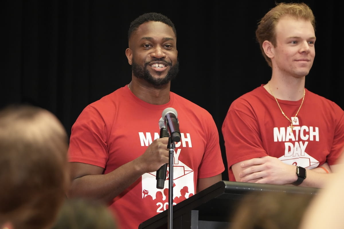 Myke Spencer holds a microphone, standing next to another medical student in Match Day Tshirts