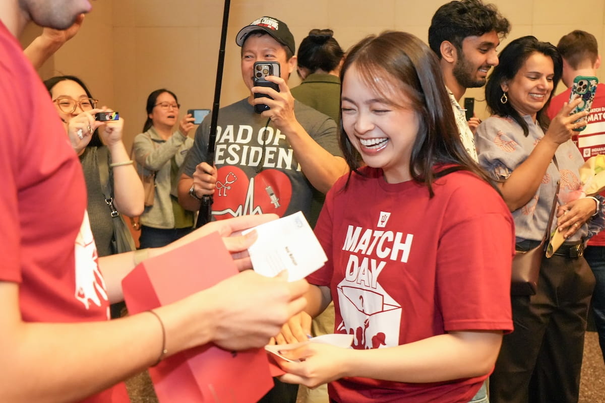 Two med students in Match Day shirts open envelopes while a man films in background