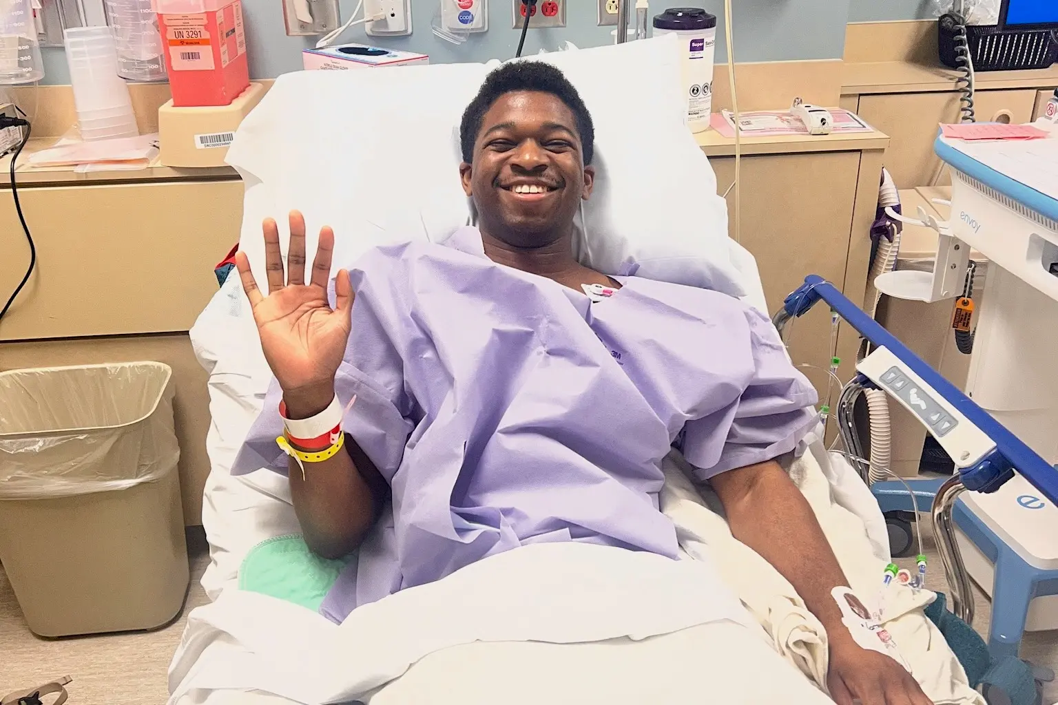 Matthew Blango lays on a hospital bed in purple gown and waves with a smile on his face
