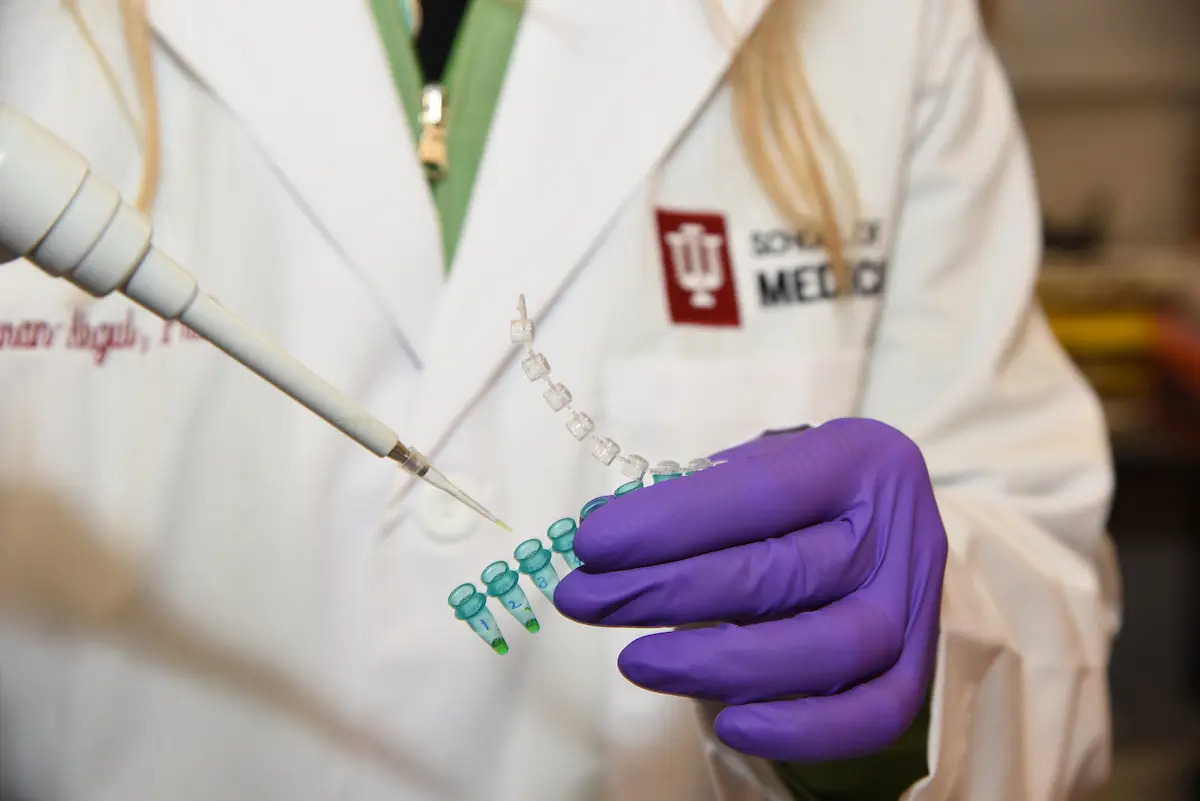 Close-up of a gloved hand in a purple nitrile glove holding a row of small vials, with a pipette dispensing liquid into one, against a white lab coat with an Indiana University School of Medicine patch.