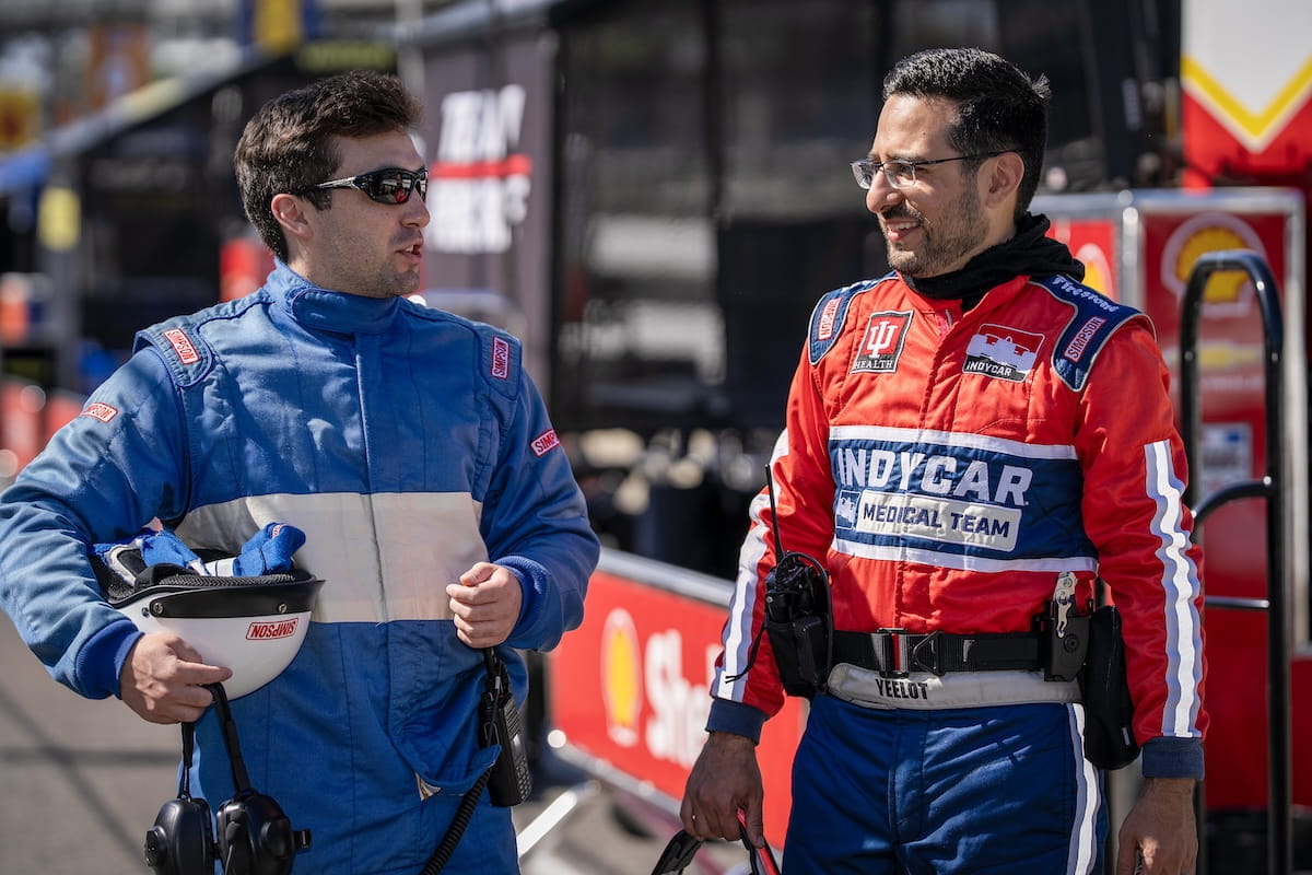 Brain Tarnai, in blue fire suit with helmet in hand, talks with Carlos Yeelot, in a red medical team fire suit, along pit lane