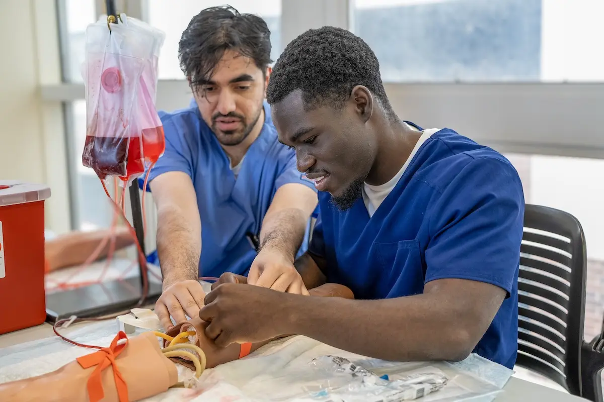 Two medical students practice starting an IV line on a manikin arm.