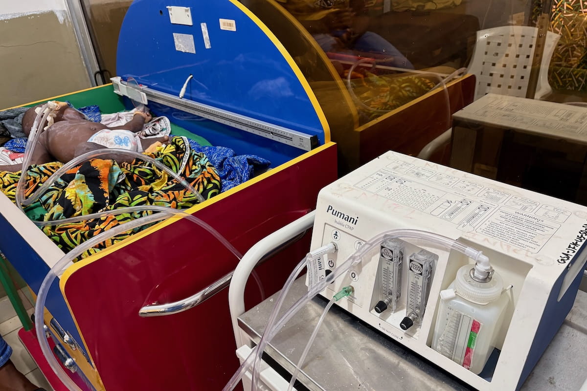 A Liberian toddler lies in a colorful hospital crib hooked up to the Pumani bubble CPAP machine.