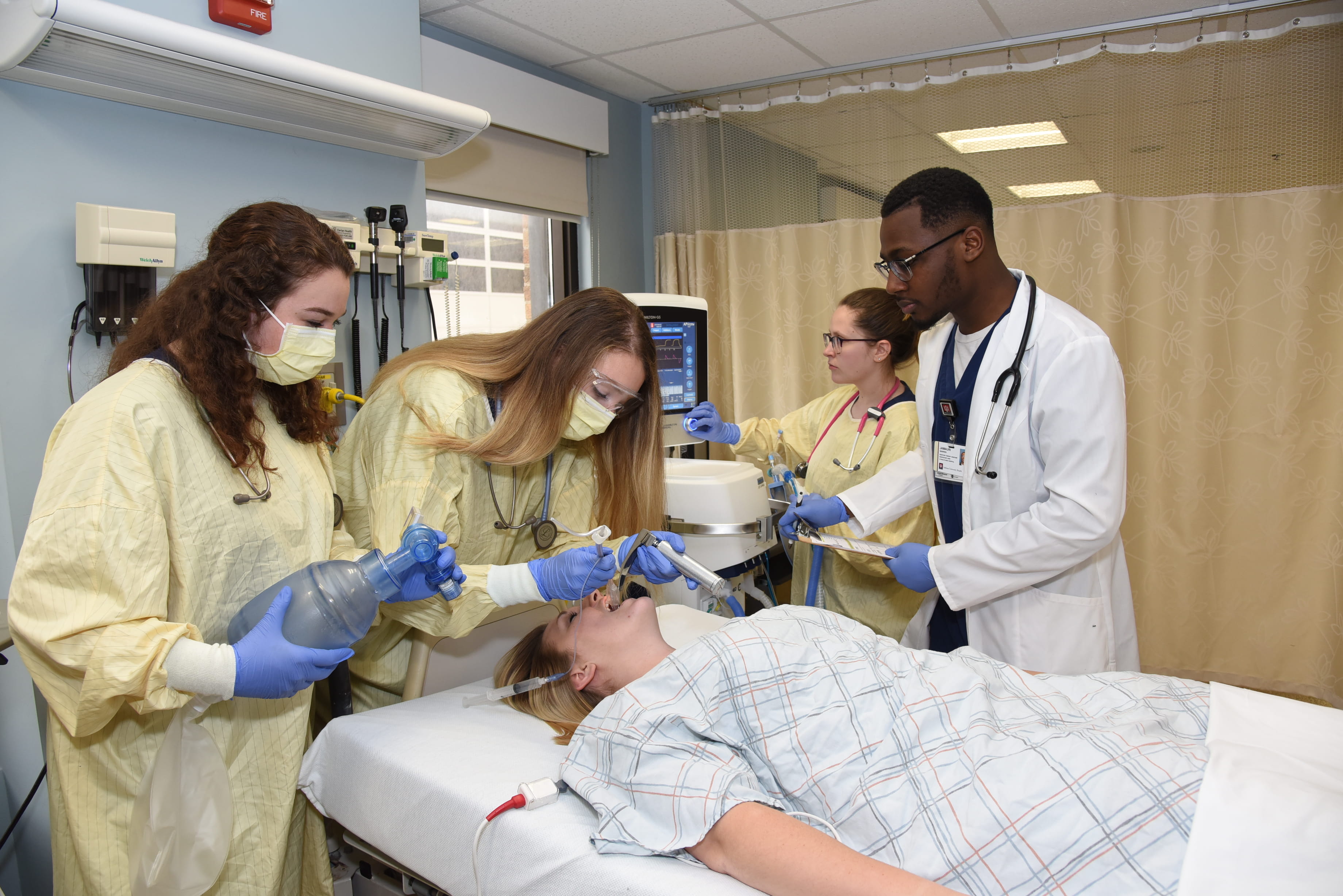 Four students in surgical scrubs practice lifesaving skills on a woman lying in a hospital bed.