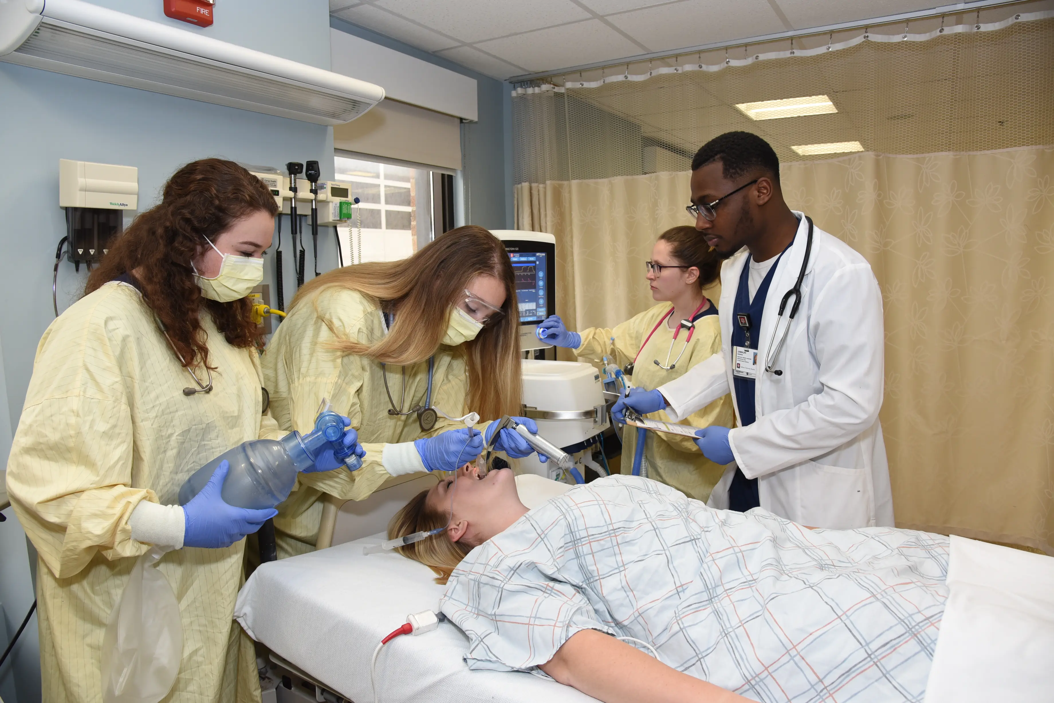 Four students in surgical scrubs practice lifesaving skills on a woman lying in a hospital bed.