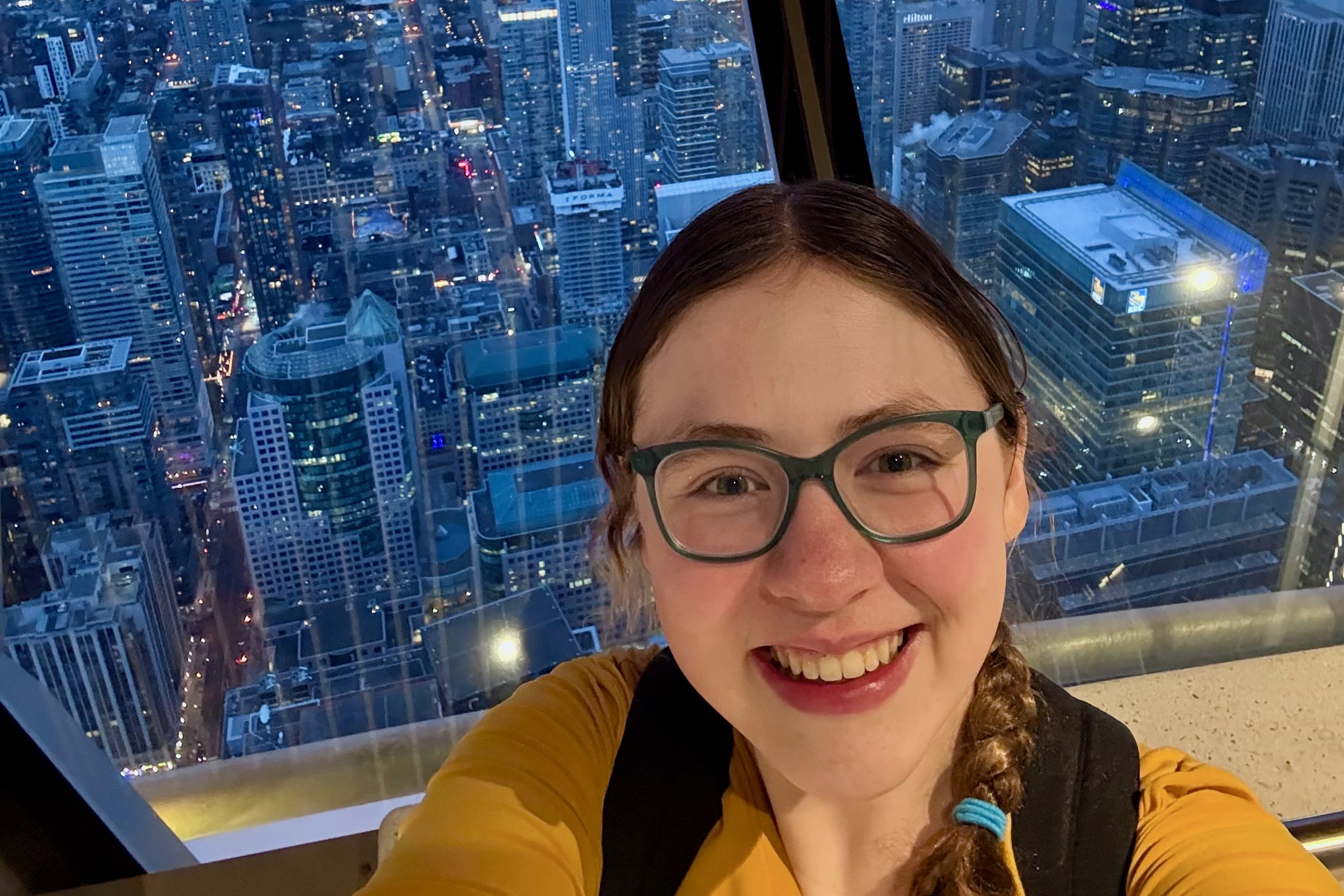 Selfie of Sarah Vaught from the top of the CN Tower in Toronto with skyscrapers behind her at night