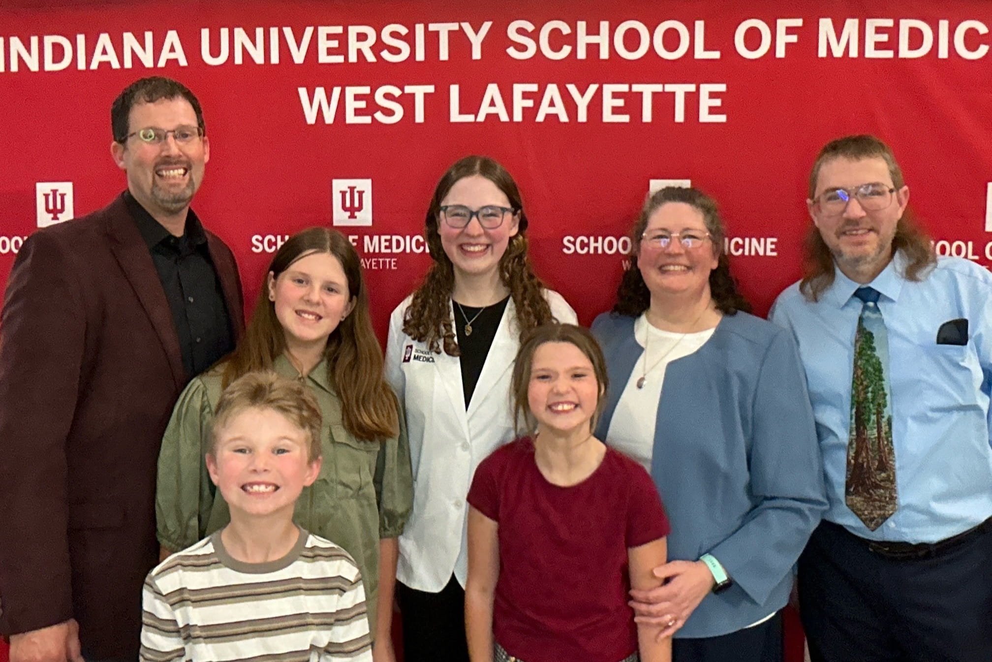 Sarah Vaught poses with her family at her white coat ceremony in front of a red IU School of Medicine—West Lafayette backdrop