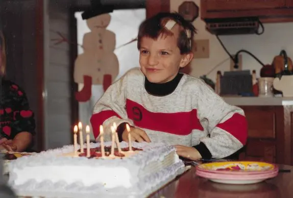 A young Kyle Gospodarek sits at a table with a cake in front of him for his seventh birthday. A surgical bandage is visible on his head.