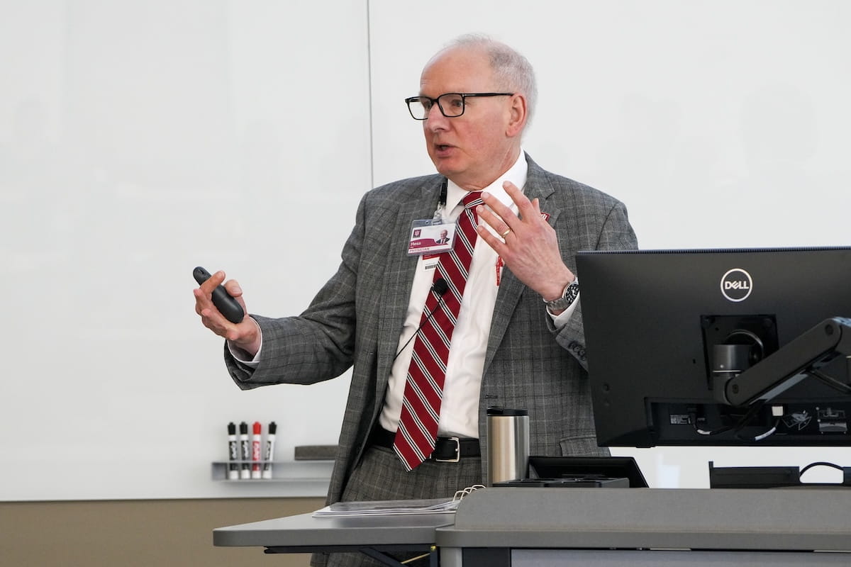 Jay Hess speaks at the front of a lecture hall, wearing suit and tie