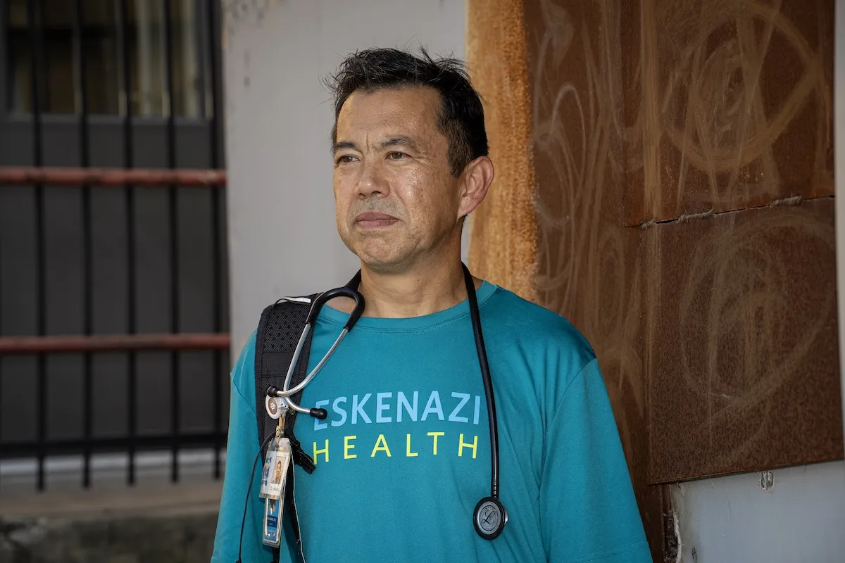 Ira Means stands by a rusted wall and looks off in the distance. He is wearing an Eskenazi Health shirt with his stethoscope around his neck. 