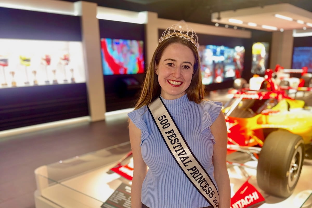 Sydney Clements wears her tiara and sash while posing in front of a race car in the museum