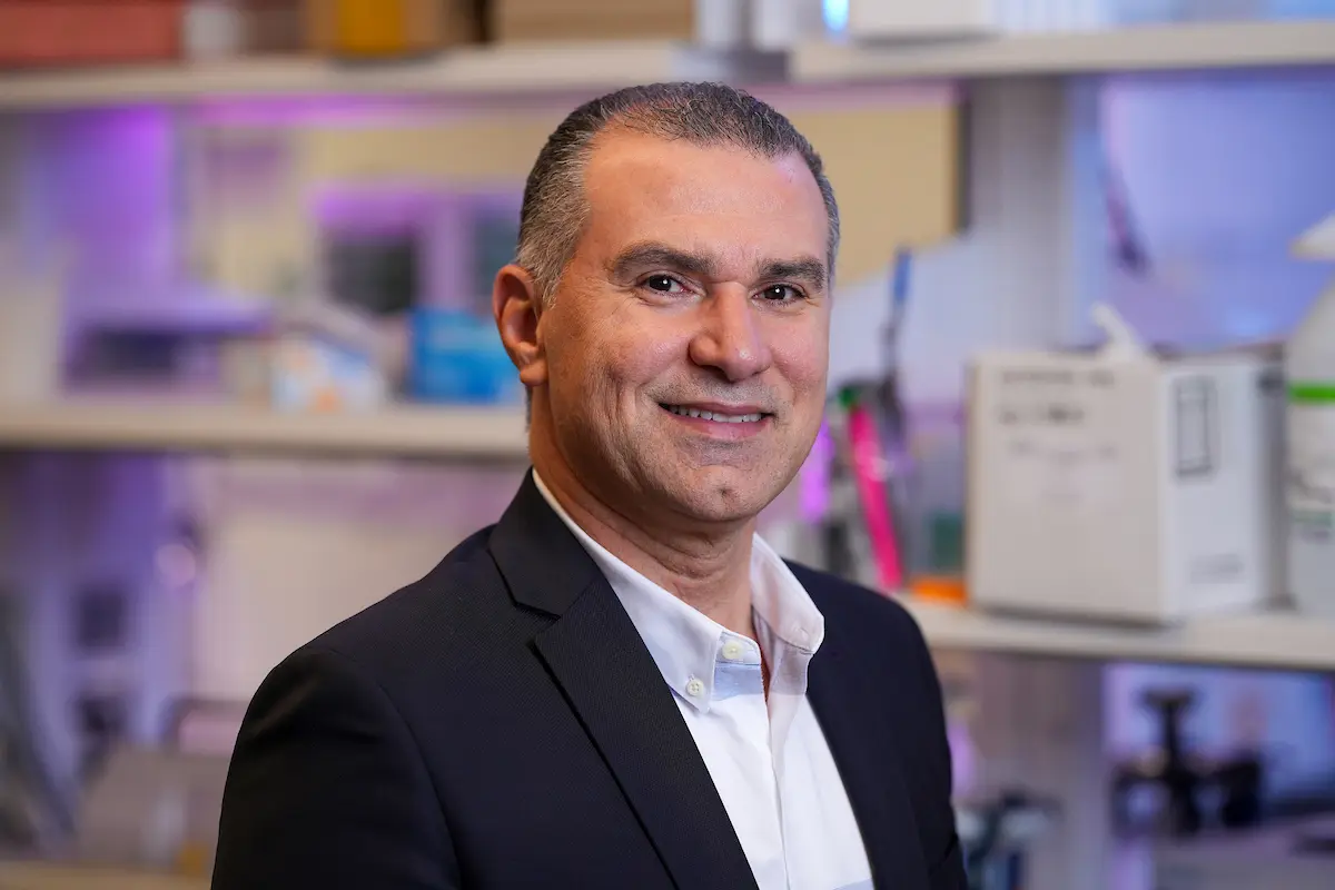 Headshot of a man with short gray hair, smiling, wearing a navy blazer over a white shirt, standing in a laboratory with shelves of equipment in the background. Photo/Image courtesy: IU School of Medicine