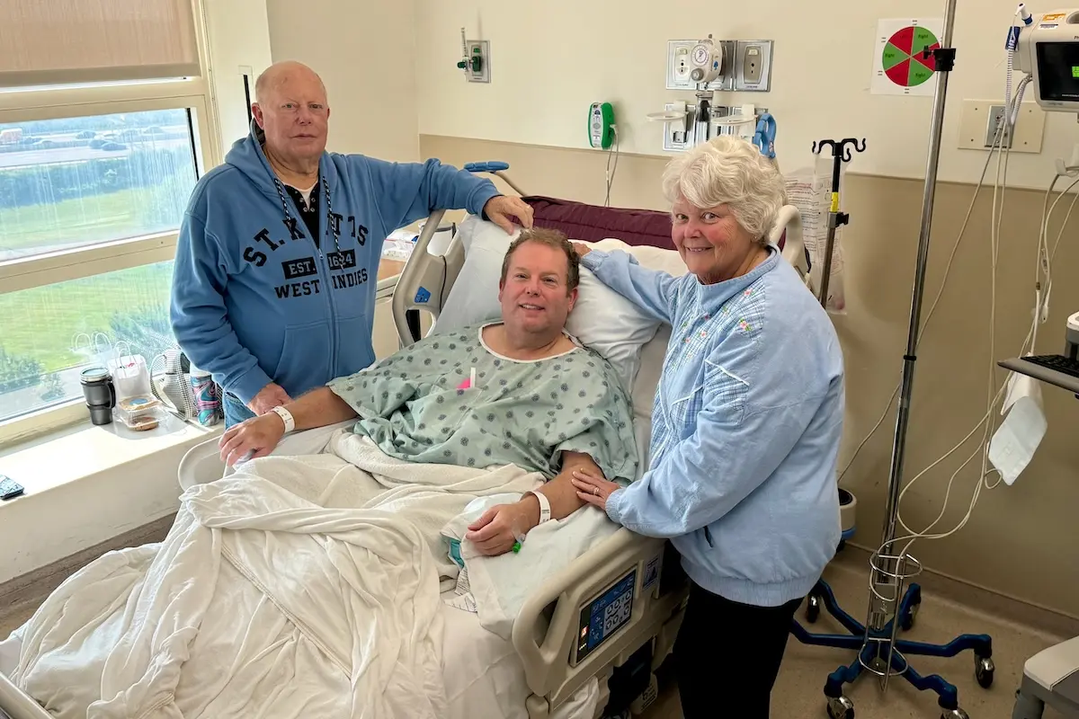 Brady Powers lies in a hospital bed with his parents standing at either side of the bed. Powers is smiling and wearing a hospital gown.