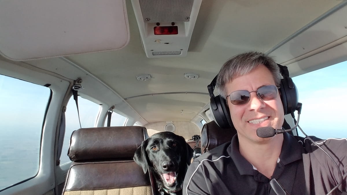 John Turchi at the pilot's wheel inside a plane with a black lab dog