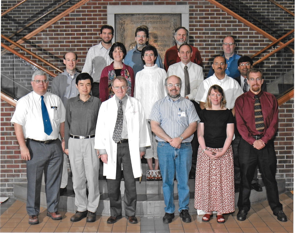 A group of 16 people in business attire pose in front of a staircase with brick wall behind them. Dr. David Weaver, in his white coat, stands at the front.