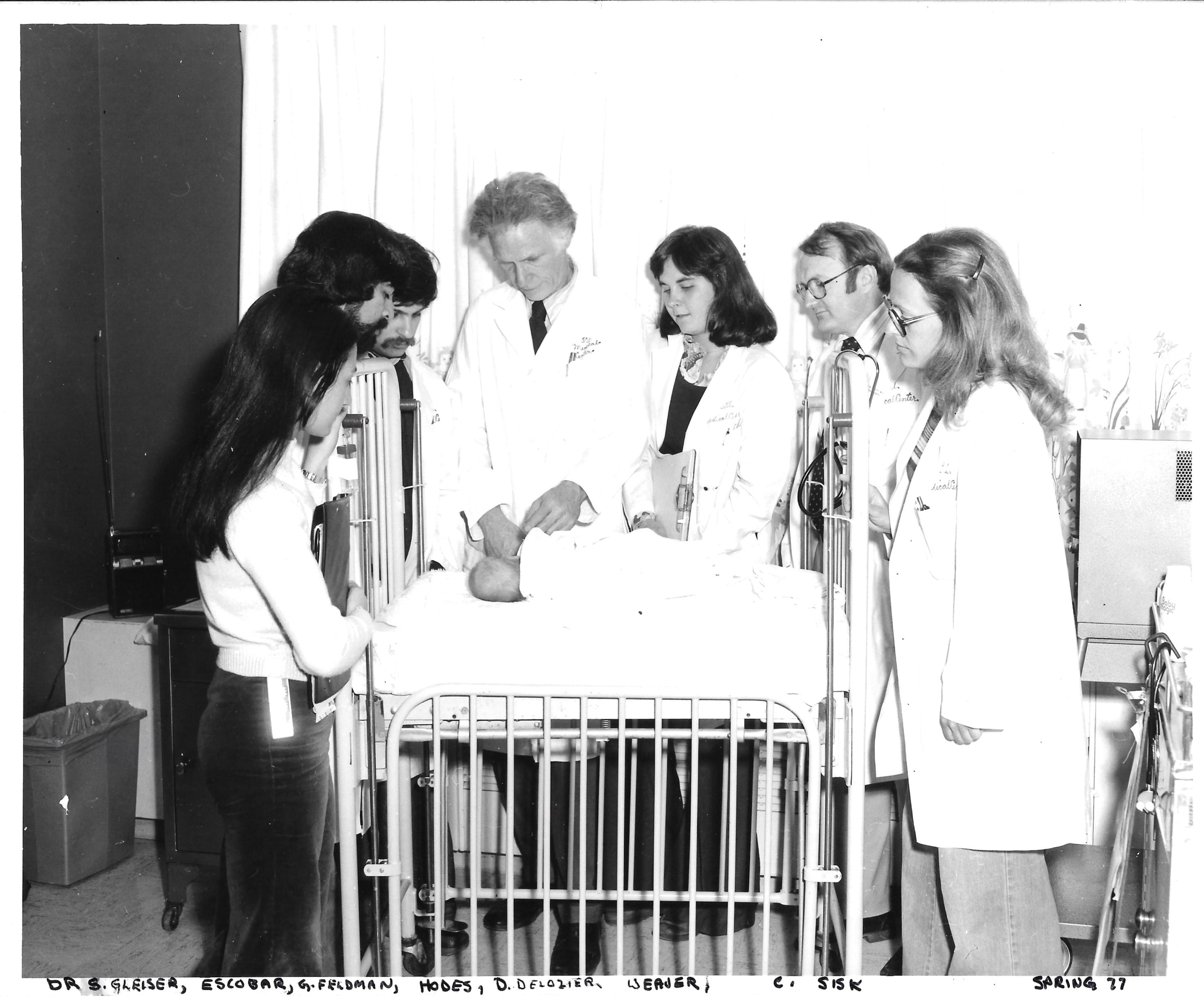 A black-and-white photo of a group of doctors wearing white coats examining an infant in a clinical setting