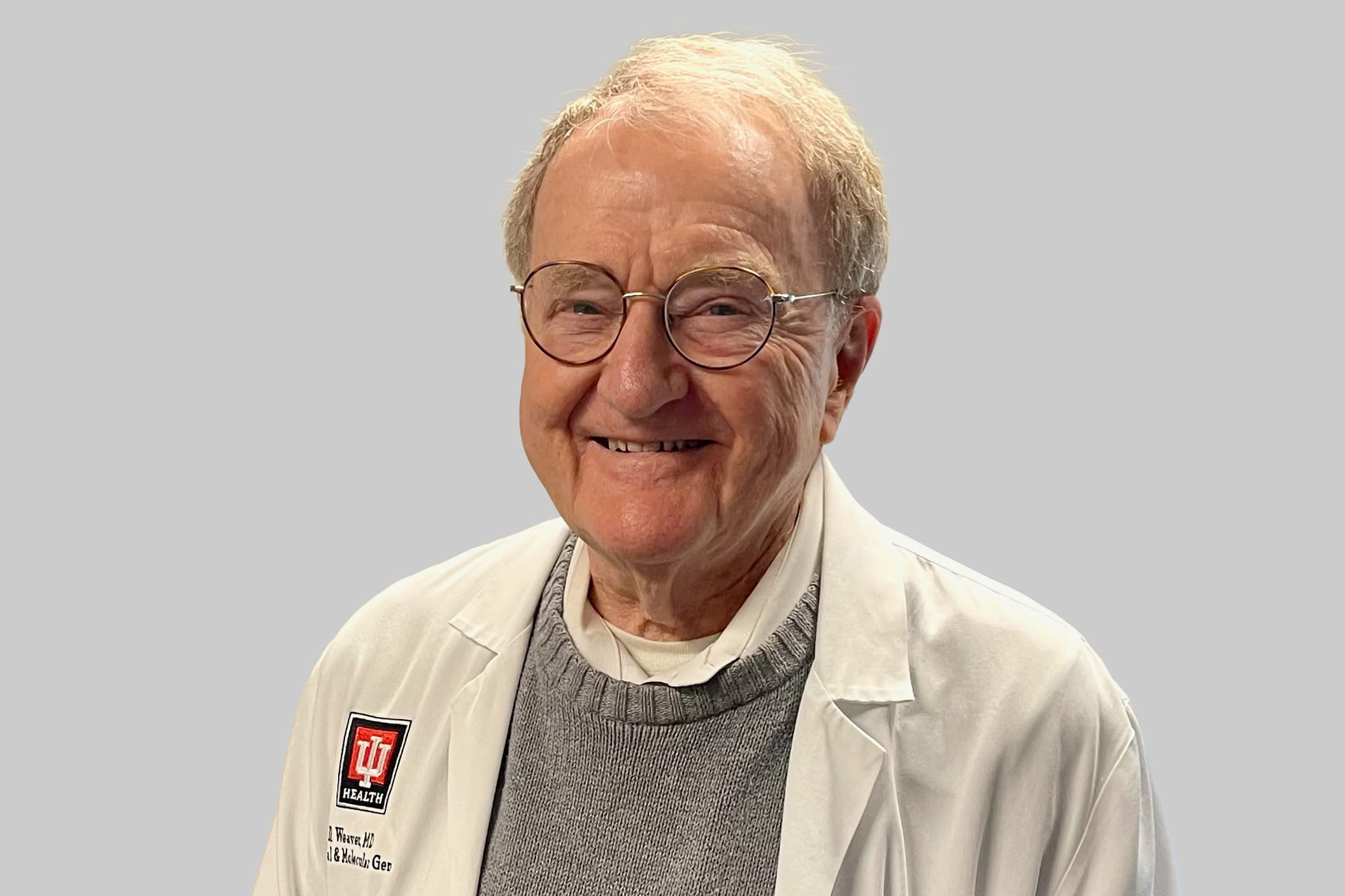 Portrait of David Weaver, older man smiling and wearing glasses, a gray sweater and an IU logo white coat; solid gray background
