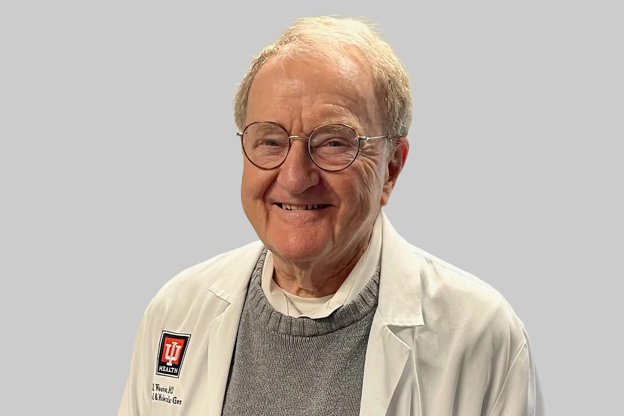 Portrait of David Weaver, older man smiling and wearing glasses, a gray sweater and an IU logo white coat; solid gray background