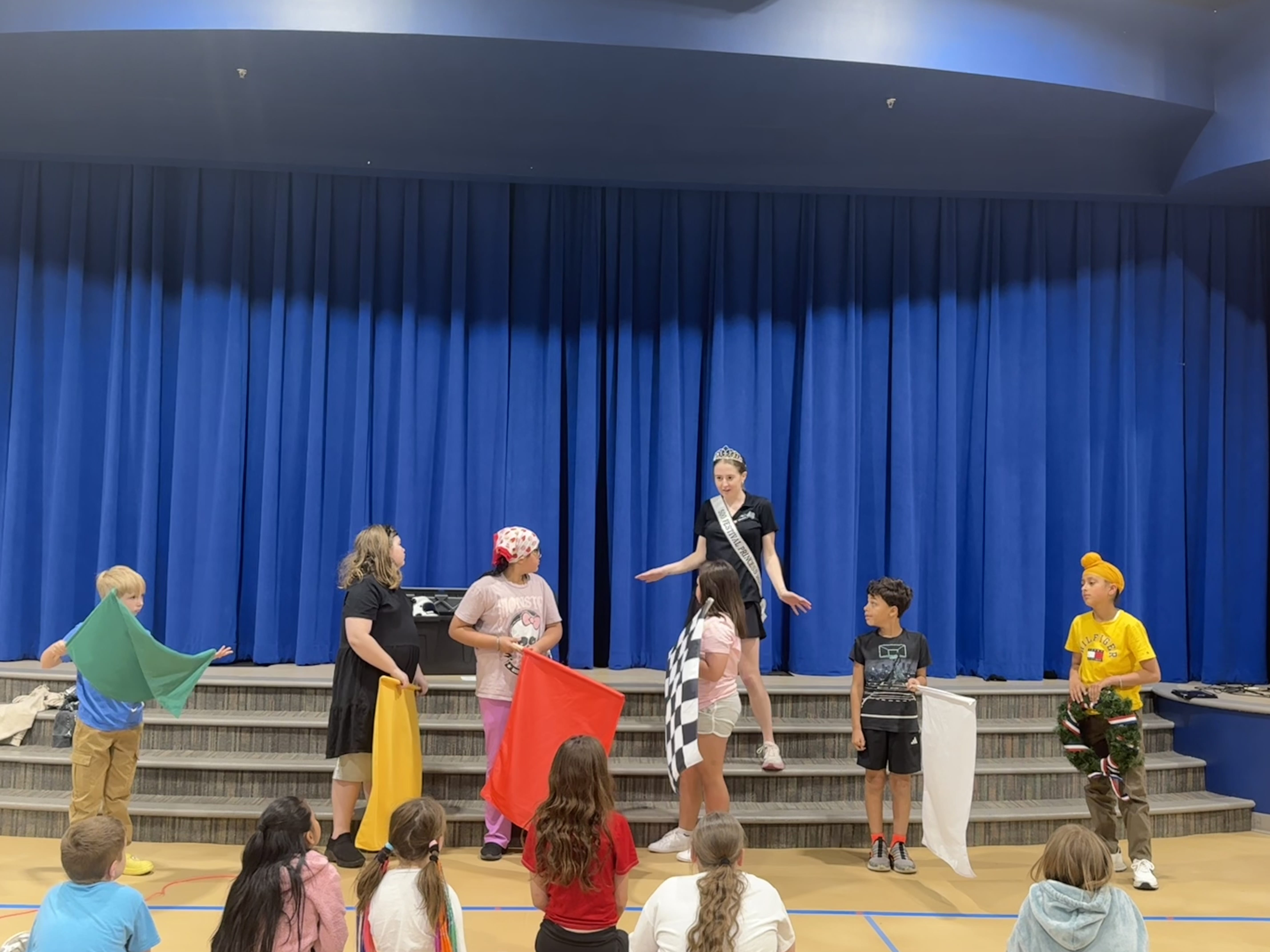 Children hold colorful flags as Sydney Clements leads a program from the stage in an elementary school gymnasium