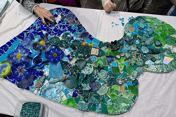 Image shows hands working on a mosaic project made up of blue and green pieces.
