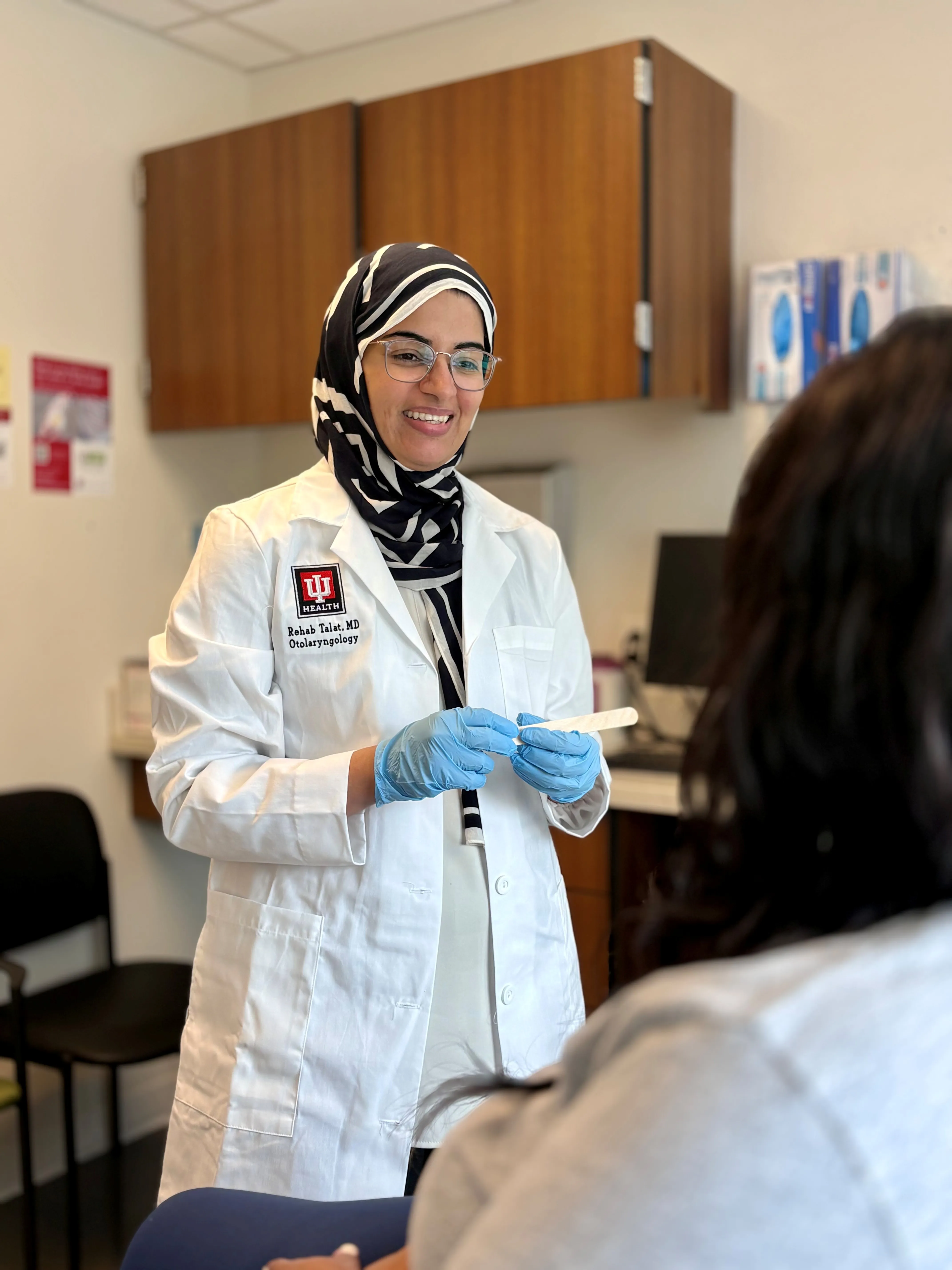 Clinician in a white coat and gloves speaks with a patient in an exam room while holding a medical swab.