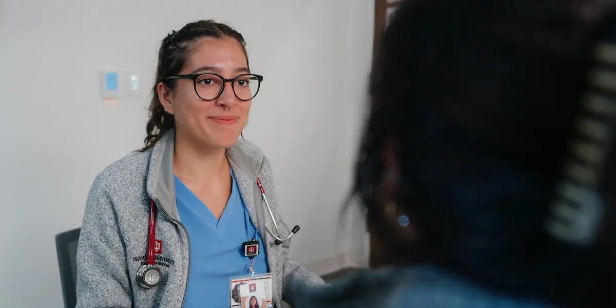 A medical student in blue scrubs smiles at a patient as they talk in the clinic.