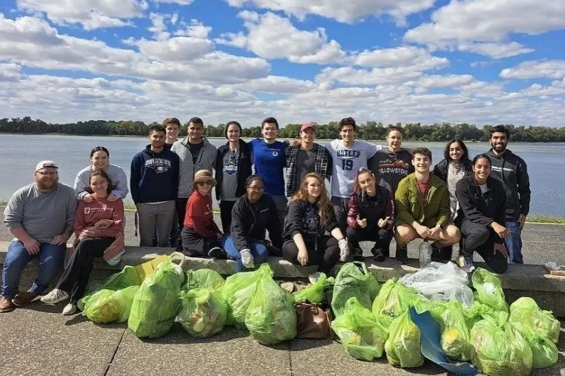 a large group of students stand with their arms around each other in front of a large river. in front of them are 25+ bags filled with trash. There is a blue sky with scatter clouds overhead.