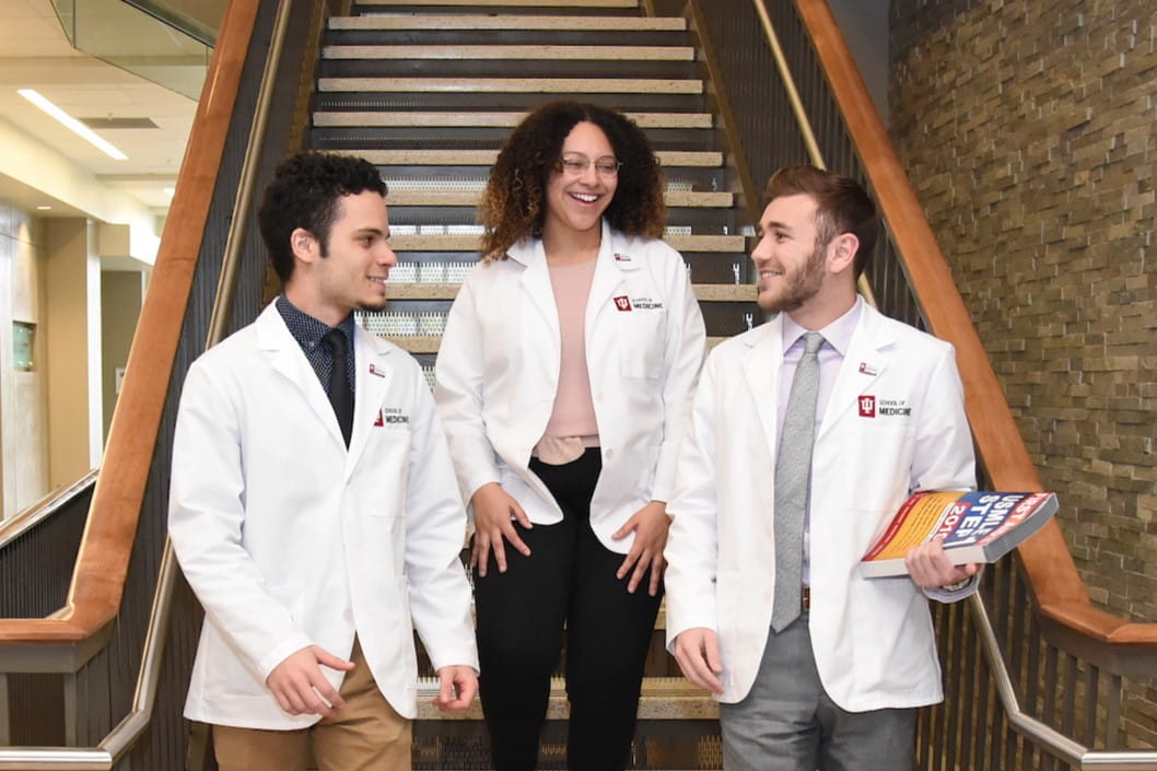 three students in white coats smiling and talking together while they walk down the stairs at the west lafayette campus.