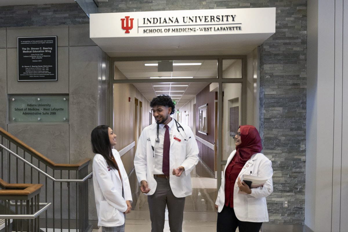 three students in white coats smile and talk together in the lobby of a building in west lafayette