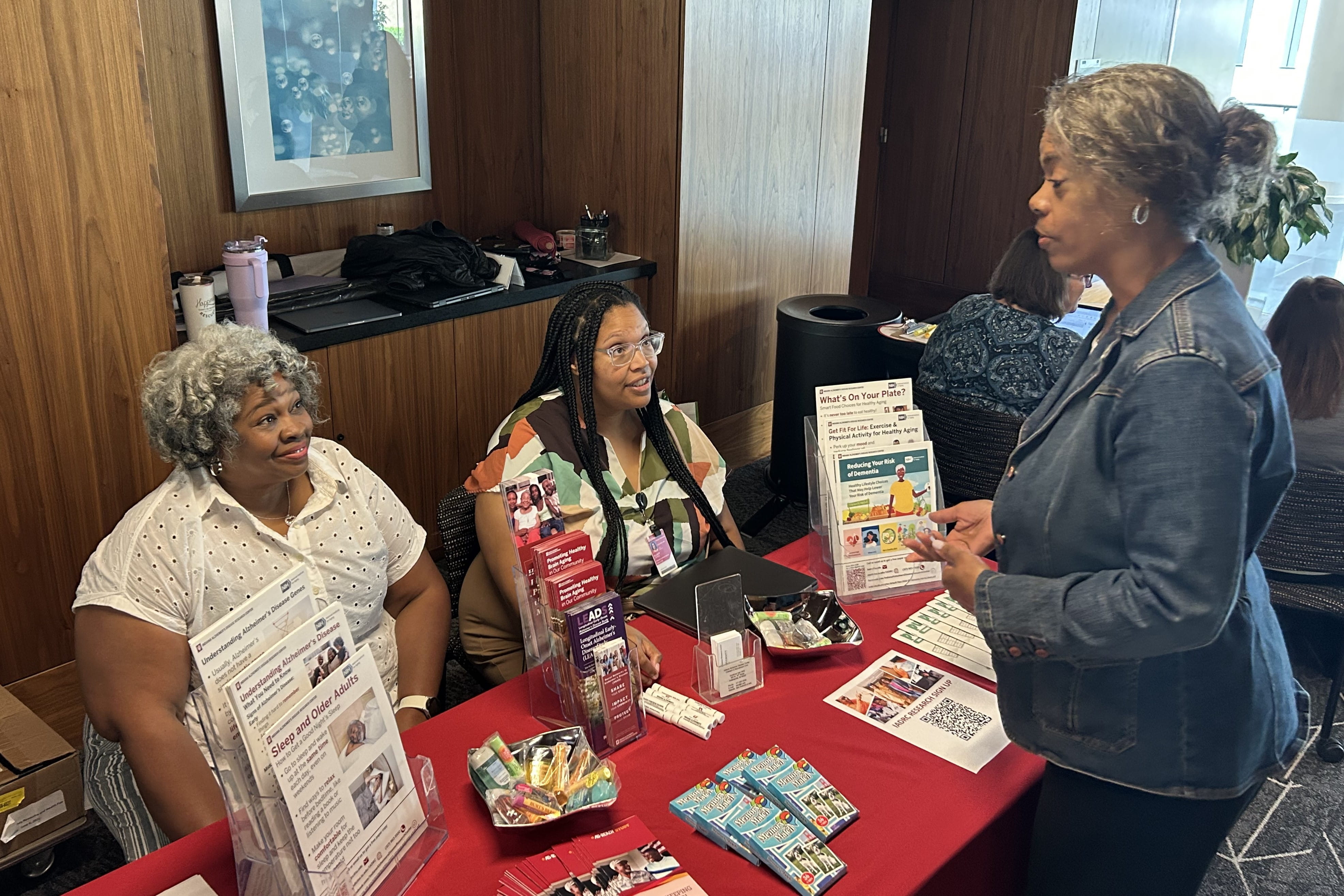 Two African-American women staff an information table covered in flyers and brochures while speaking with a community member.