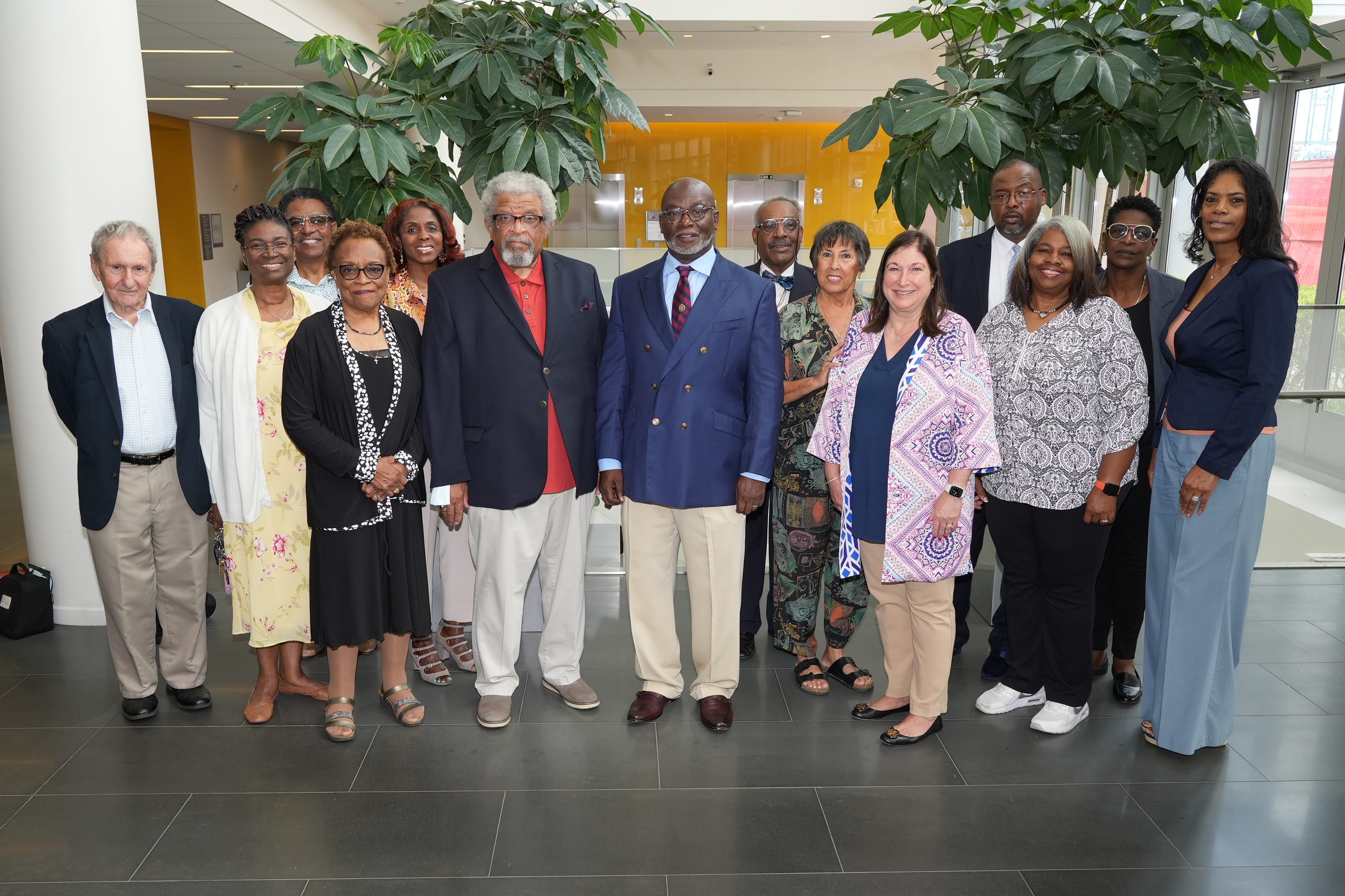 Fourteen community advisory board members stand together in a building lobby with windows and plants in the background.