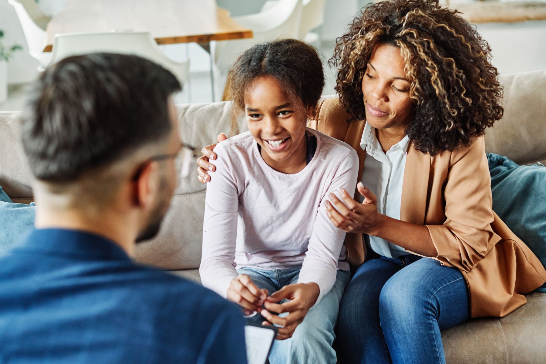 a child and parent sit close together on a couch and smile towards a mental health provider