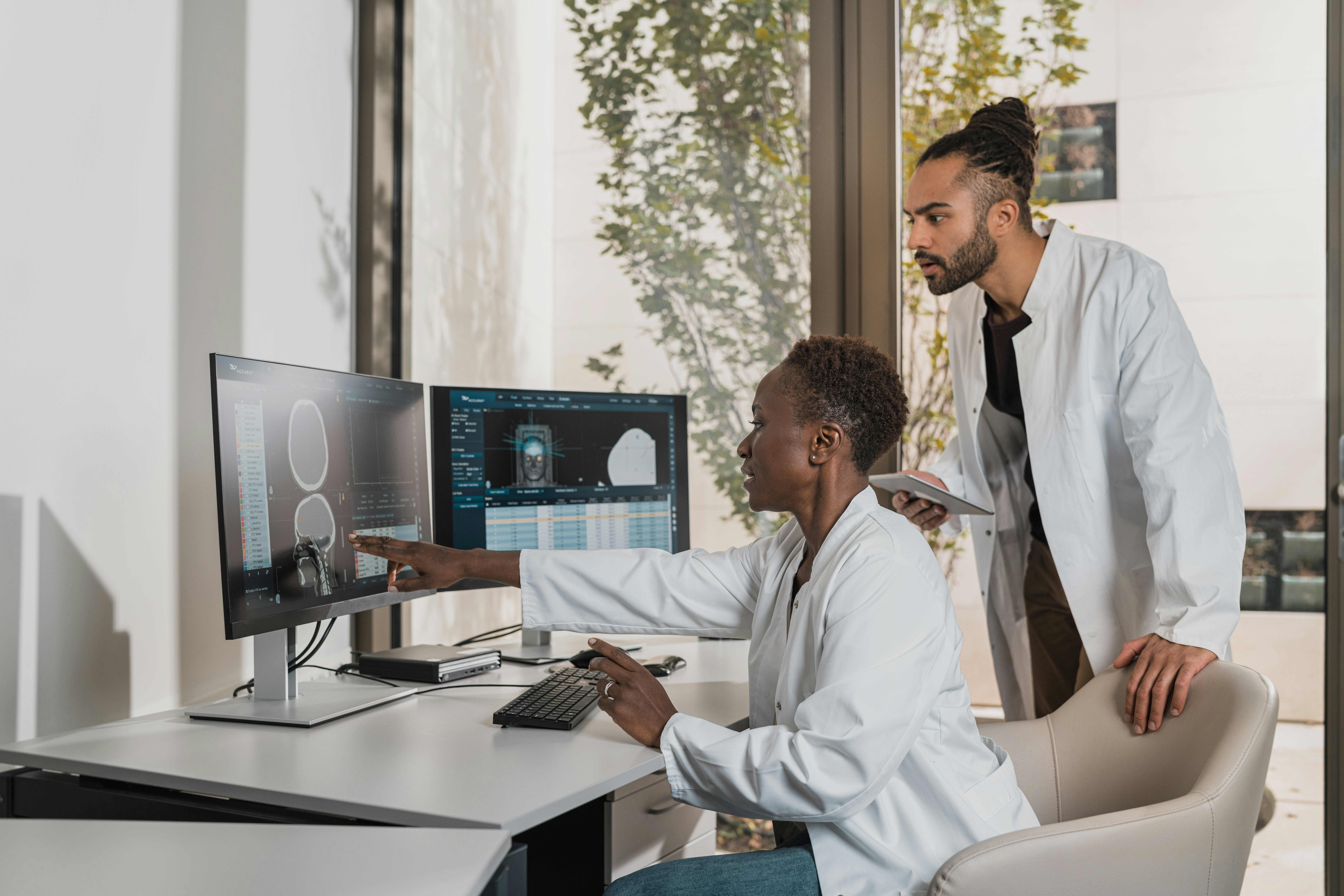 Two medical professionals in lab coats review medical imaging on a computer screen, collaborating in a bright office with large windows and a view of trees outside.