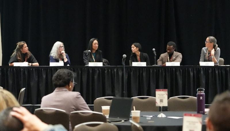 six panelists of diverse backgrounds sit at a long table in the front of a conference room