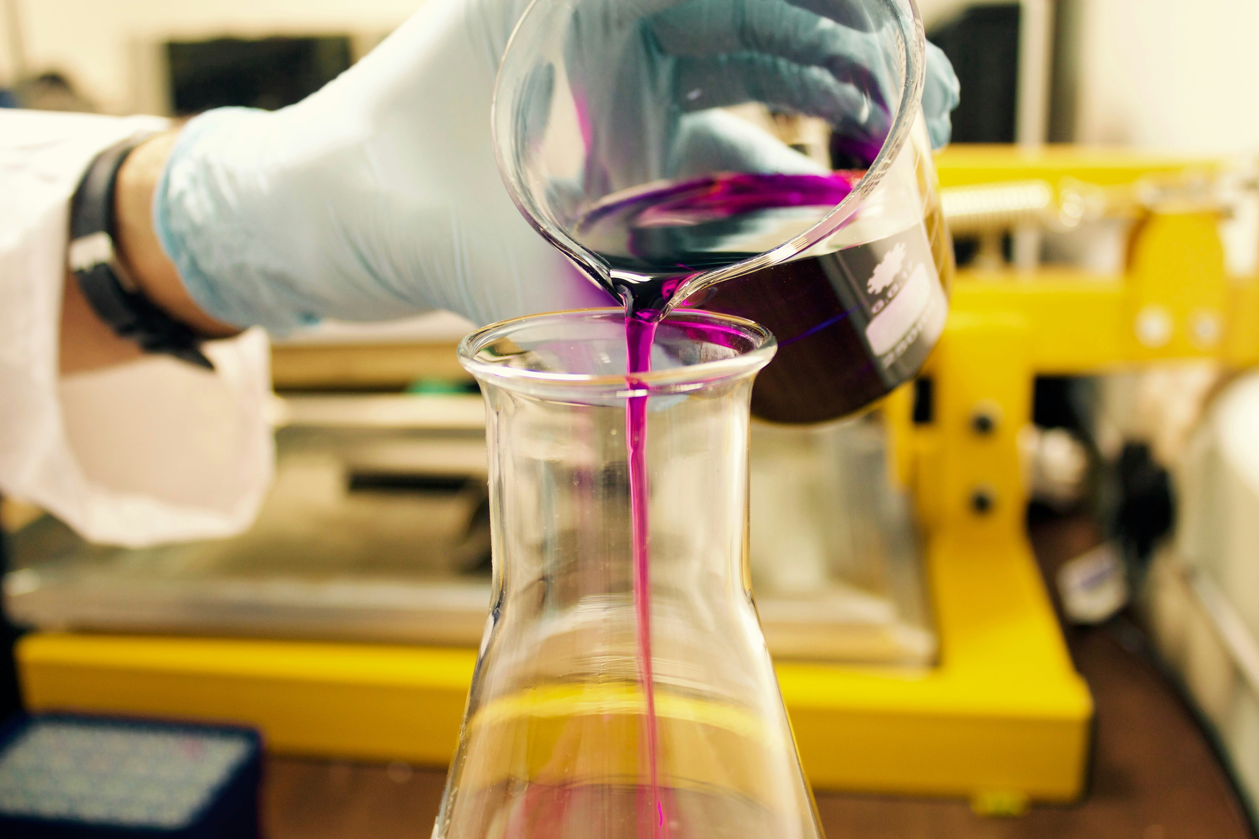 A gloved hand pours a vivid purple liquid from a beaker into a glass flask in a laboratory, with blurred yellow equipment in the background.