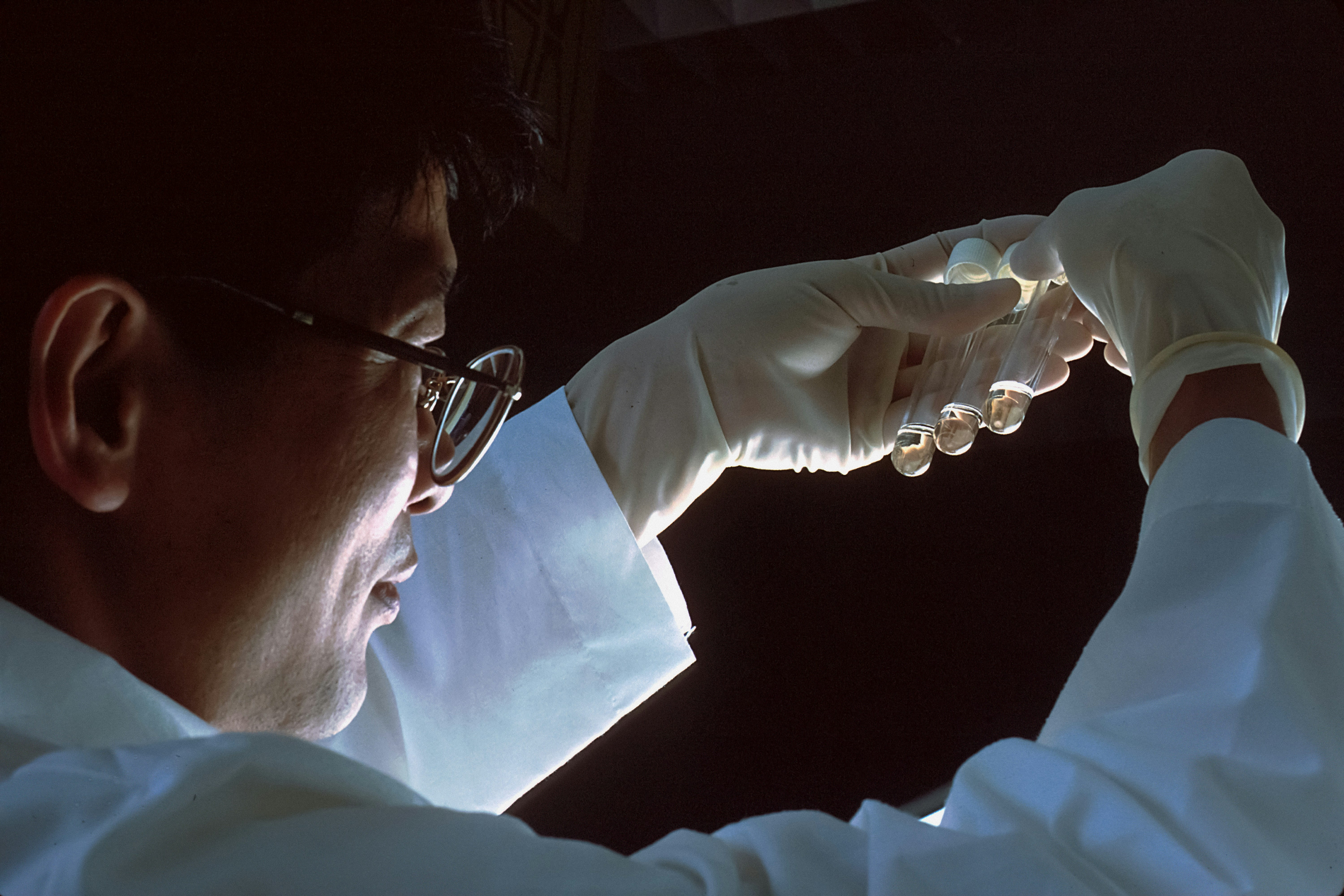 A scientist in a lab coat and gloves examines test tubes under focused lighting, highlighting the contents against a dark background.