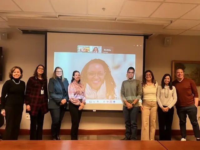 Graduation ceremony for HERS Scholars: A group of people stands together in front of a projector screen displaying virtual attendees on Zoom, celebrating the scholars' achievements.