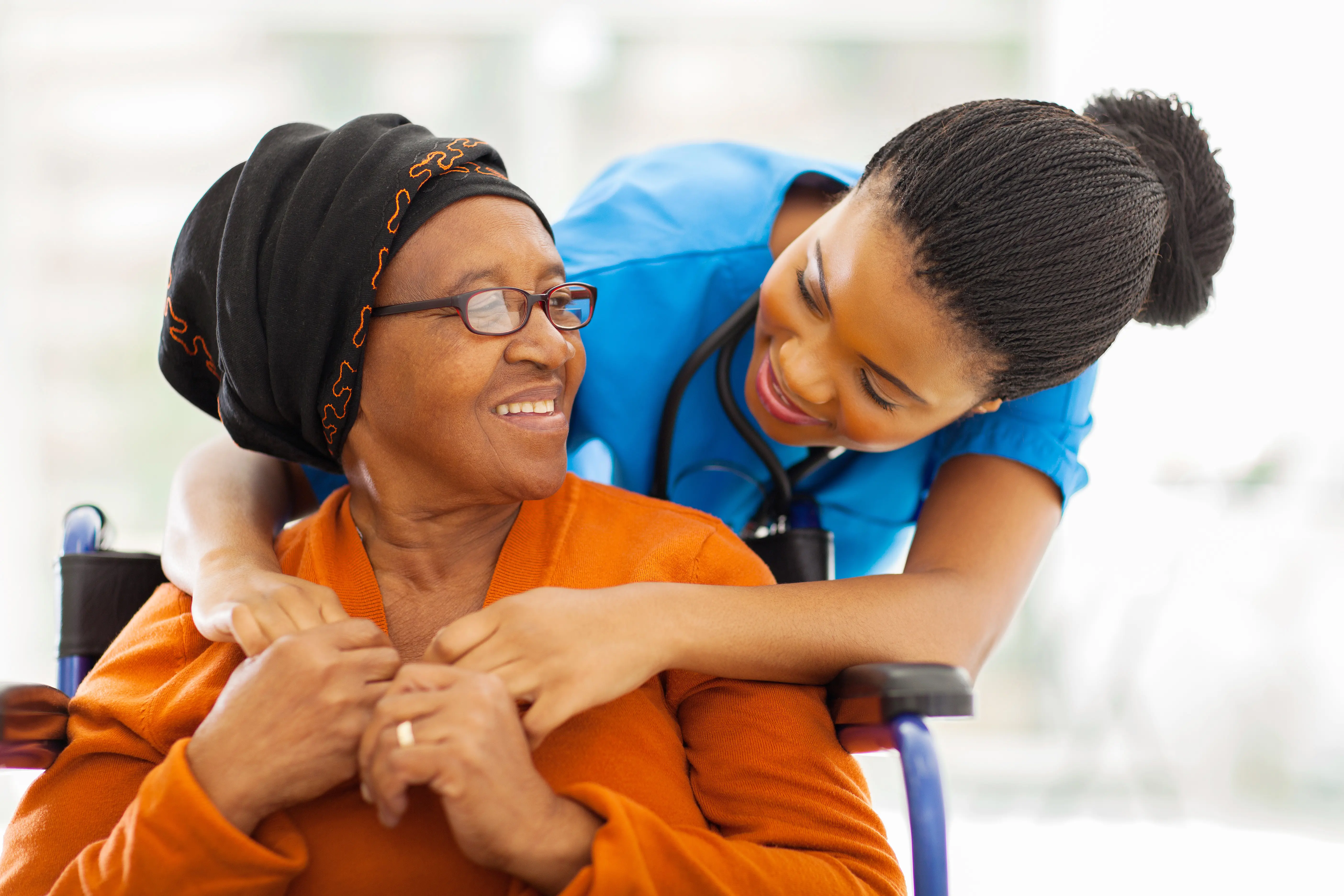 A health care worker in blue scrubs leans over with her arms around an older woman in a wheelchair