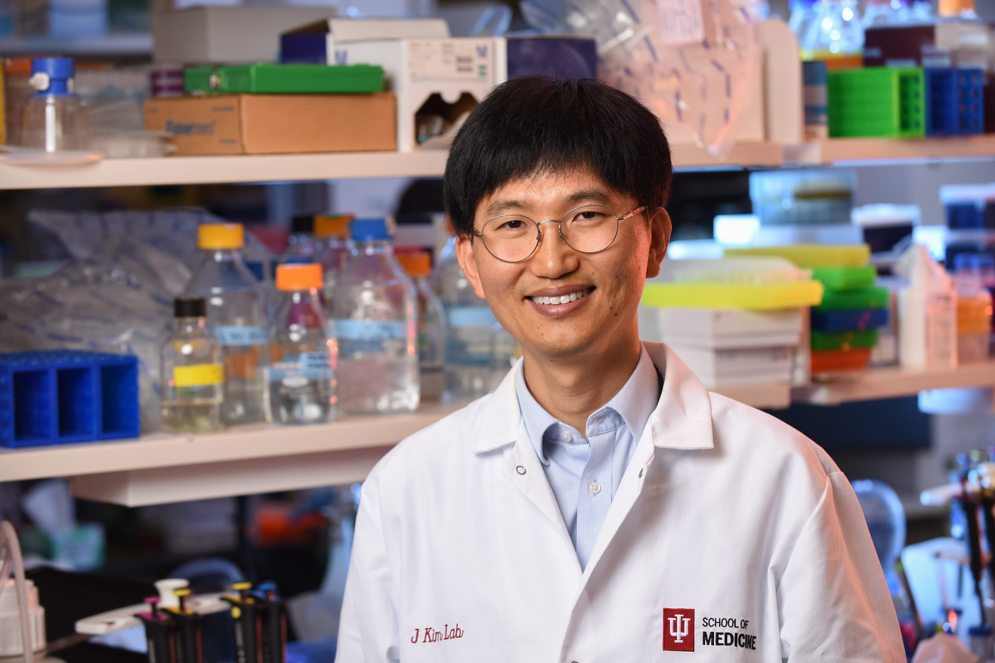 A portrait of Jungsu Kim in a lab, wearing an IU School of Medicine branded white coat.