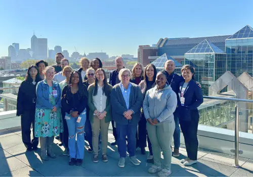 Group photo of 17 Gaston Lab group members posed outside with IU School of Medicine campus and Indianapolis skyline in the background