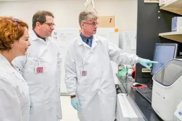 Three researchers in white coats look at an instrument in the lab