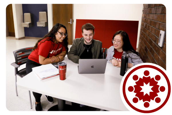 Three college students sit together at a table while working. One student works on a laptop while the others lean in collaboratively.