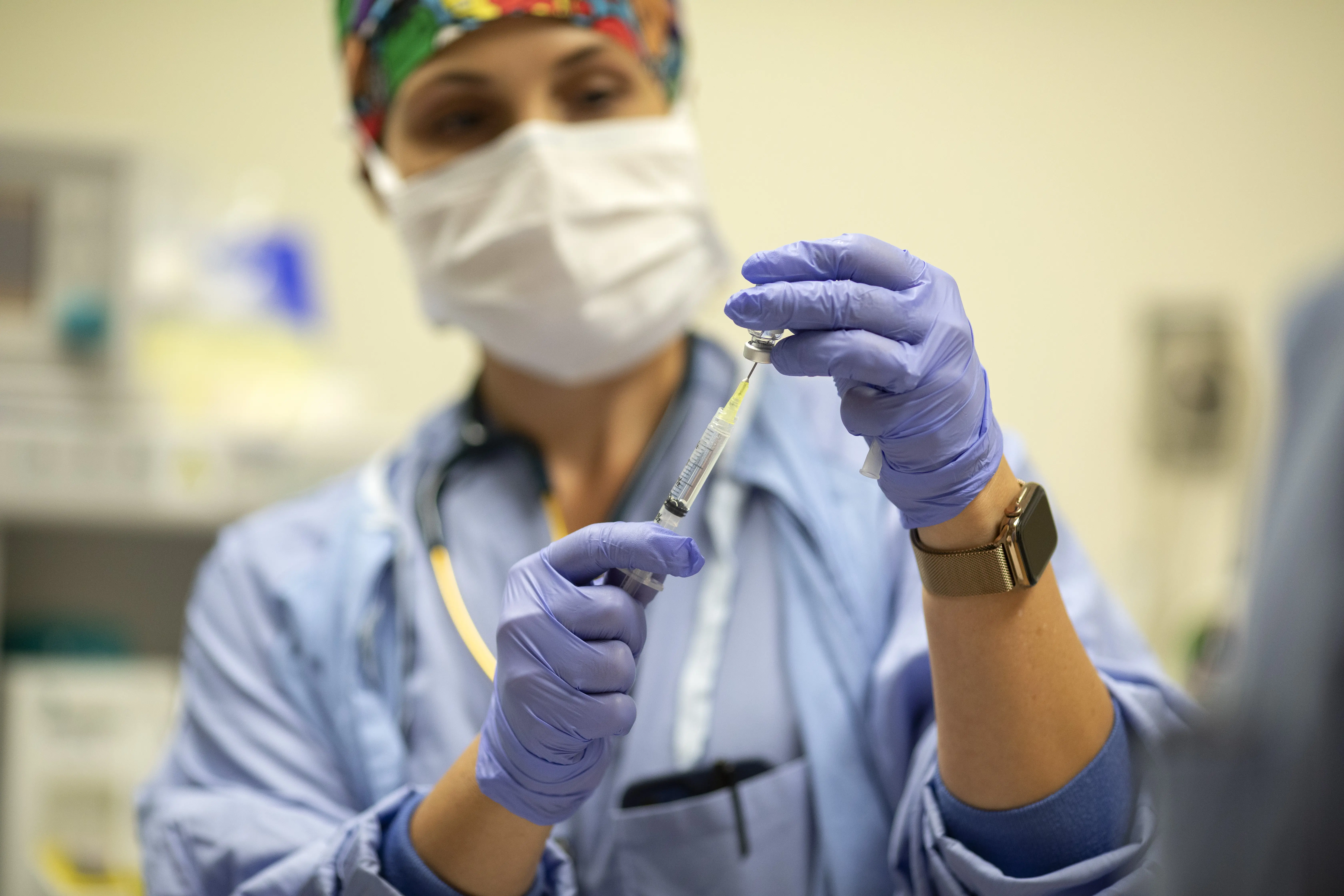 Masked clinician wearing gloves prepares medication in a syringe in a clinical setting.