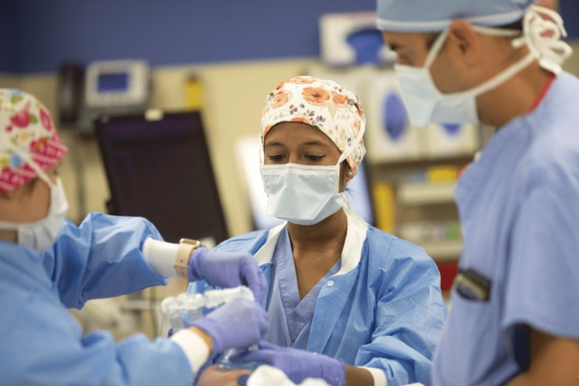 Medical team in surgical attire placing an airway mask on a patient in an operating room.